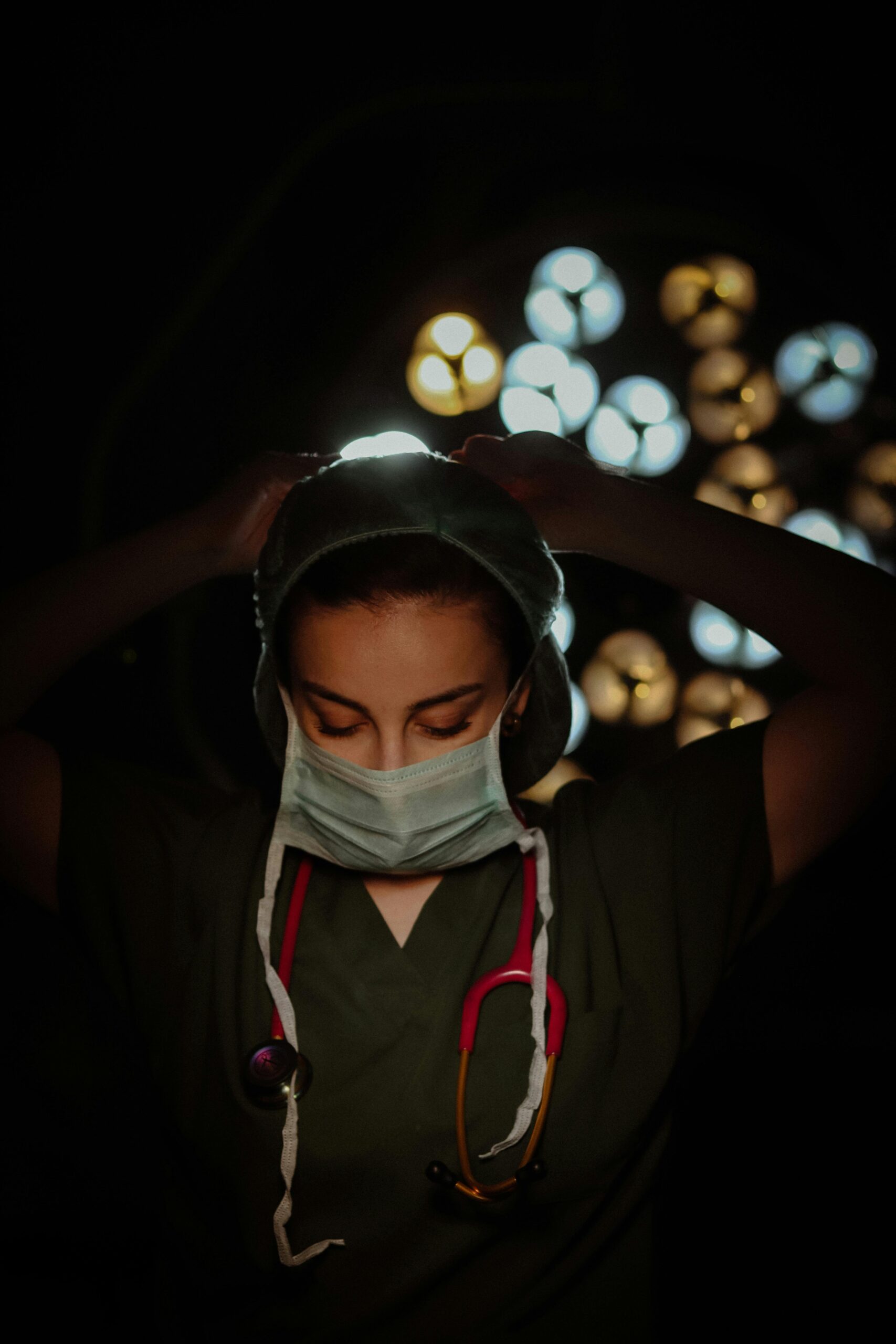 A female surgeon adjusts her mask under surgical lights, ready for operation.